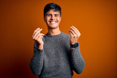 İzole edilmiş turuncu arka plan üzerinde duran genç ve yakışıklı bir adam. Elleriyle para hareketi yapıyor. Maaş ödemesi istiyor.