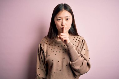 Genç, güzel Asyalı bir kadın pembe arka planda şık bir süveter giyiyor ve parmakları dudaklarında sessiz olmak istiyor. Sessizlik ve gizli konsept.
