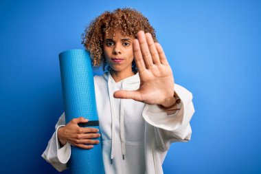 Genç Afro-Amerikalı kadın, yoga minderini kapalı mavi arka planda tutarak spor yapıyor. Açık elle, ciddi ve kendinden emin bir ifadeyle dur işareti yapıyor.