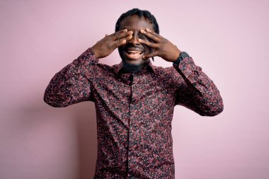 Genç, yakışıklı, Afro-Amerikan bir adam, günlük gömleğini giymiş pembe arka planda dikiliyor, gözleri neşeli ve komik bir şekilde gülümsüyor. Kör kavram.