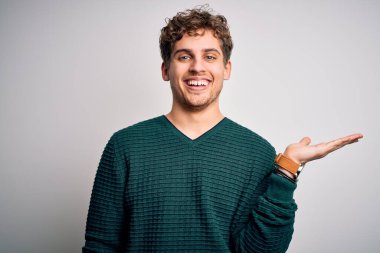 Sarışın, kıvırcık saçlı, yeşil süveter giyen, beyaz arka planda gülümseyen, eli kameraya bakan genç ve yakışıklı bir adam..
