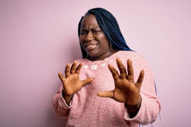 Afro-amerikan kadın örgü örüyor pembe arka planda günlük süveter giyiyor tiksindirici ve tiksindirici bir yüz ifadesi takınıyor çünkü tiksinti tepki veriyor. Eller havada