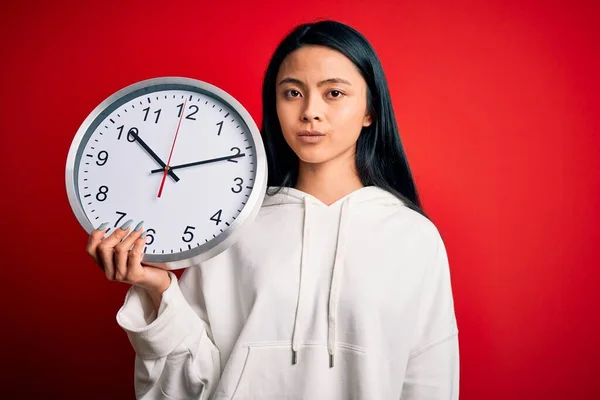 年轻美丽的中国女人 拿着大钟 站在孤零零的红色背景上 脸上带着自信的表情 严肃地思考着 — 图库照片