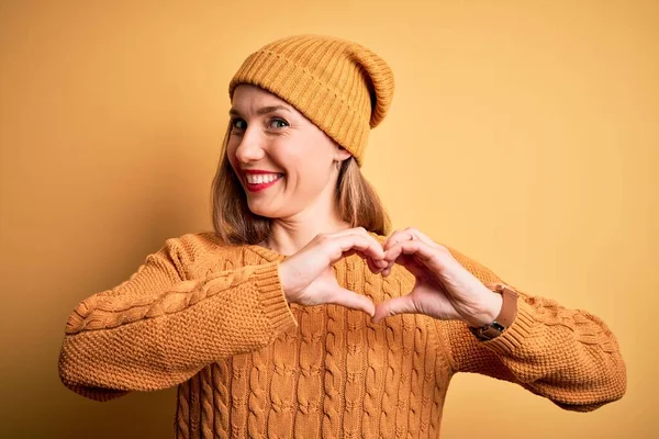 黄色の背景にカジュアルなセーターとウールキャップを身に着けている若い美しいブロンドの女性は ハートシンボルと手で形を示す愛に笑みを浮かべています ロマン主義 — ストック写真