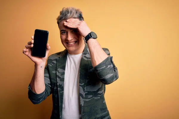 Young modern man showing smartphone blank screen over yellow isolated background stressed with hand on head, shocked with shame and surprise face, angry and frustrated. Fear and upset for mistake.