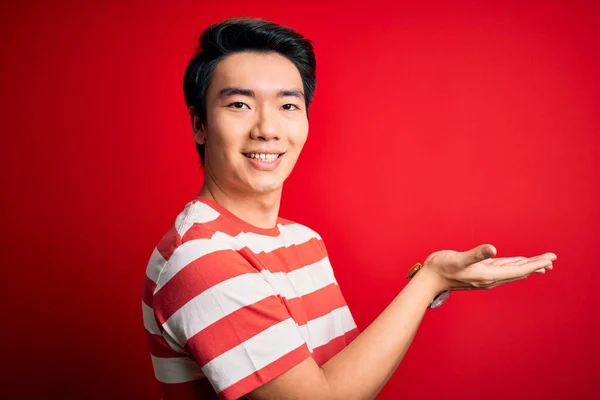 若いハンサムな中国人男性を着てカジュアルな縞模様のTシャツ赤い背景の上に立って脇に手を開いてヤシの木は コピースペースを示す 広告笑顔興奮幸せな提示 — ストック写真