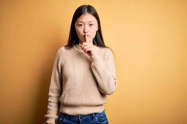 年轻美丽的亚洲女人穿着宽松的毛衣 披着黄色孤立的背景 要求保持安静 手指放在嘴唇上 沉默和秘密概念 — 图库照片