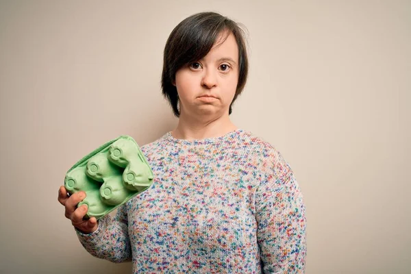 Young down syndrome woman holding cardboard egg cup from fresh healthy eggs with a confident expression on smart face thinking serious