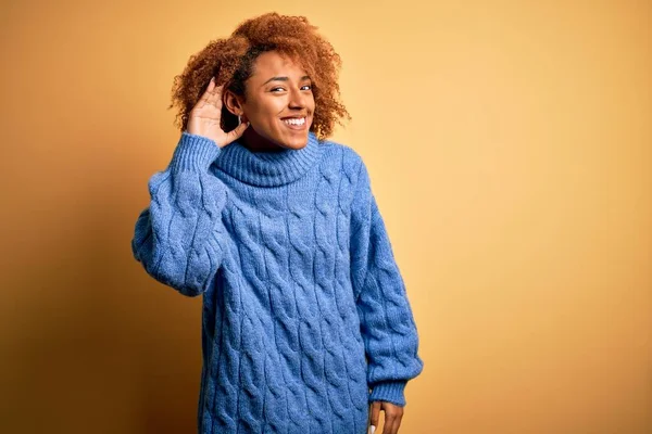 Young beautiful African American afro woman with curly hair wearing blue turtleneck sweater smiling with hand over ear listening an hearing to rumor or gossip. Deafness concept.