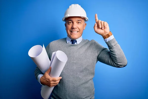 中年英俊的白发苍苍的建筑师 头戴安全帽 拿着设计图 面带微笑 用手指和胳膊指指着 — 图库照片