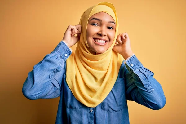 黄色の背景にアラブの伝統的なヒジャーブを身に着けている巻き毛を持つ若い美しい女性指で耳を引く笑みを浮かべて 面白いジェスチャー オーディション問題 — ストック写真