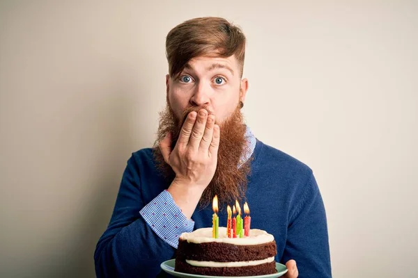Irish redhead man with beard holding birthday cake with burning candles over isolated background cover mouth with hand shocked with shame for mistake, expression of fear, scared in silence, secret concept