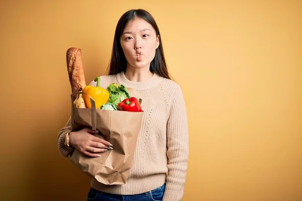 年轻的亚洲女人拿着一纸袋新鲜健康的食品放在黄色孤立的背景上 用嘴唇 疯狂和滑稽的姿势做着鱼脸 有趣的表达方式 — 图库照片