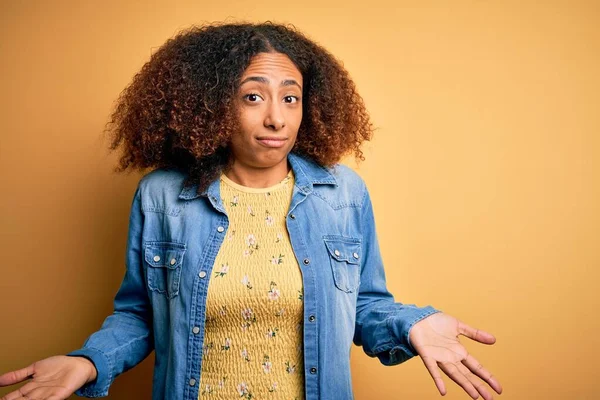 黄色の背景の上にカジュアルなデニムシャツを着てアフロの髪を持つ若いアフリカ系アメリカ人女性は 腕や手で提起と混乱表現 疑いの概念 — ストック写真