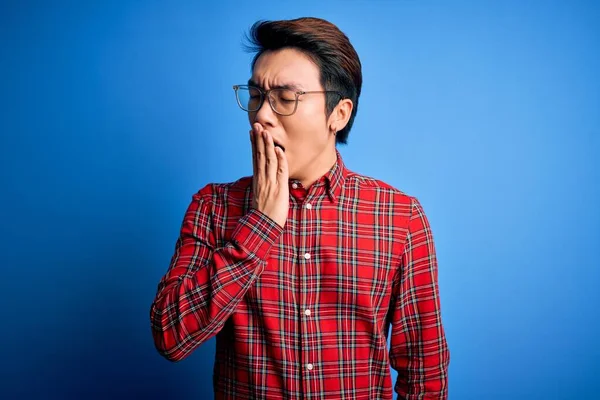 Young handsome chinese man wearing casual shirt and glasses over blue background bored yawning tired covering mouth with hand. Restless and sleepiness.