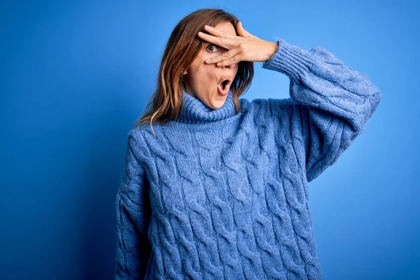 Young beautiful brunette woman wearing casual turtleneck sweater over blue background peeking in shock covering face and eyes with hand, looking through fingers with embarrassed expression.