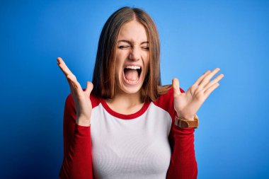 Young beautiful redhead woman wearing casual t-shirt over isolated blue background celebrating mad and crazy for success with arms raised and closed eyes screaming excited. Winner concept clipart