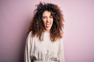 Kıvırcık saçlı ve piercingli güzel bir kadın pembe arka planda sıradan bir süveter giyiyor ve komik bir ifadeyle dilini dışarı çıkarıyor. Duygu kavramı.