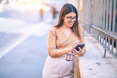 Mutlu ve kendinden emin gülümseyen genç ve güzel bir kadın. Yüzünde gülümsemeyle şehirde akıllı telefon kullanarak durmak.