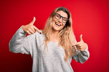 İzole edilmiş kırmızı arka plan üzerinde süveter ve gözlük takan genç, sarışın bir kadın. Kendine güvenen, yüzünde gülümseme olan, kendini gururlu ve mutlu parmaklarıyla gösteren bir kadın..