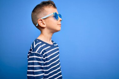 Mavi gözlü, mavi arka planda güneş gözlüğü takmış, rahat bir görünüm takınan, kendine güveni tam bir gülümsemeyle poz veren küçük beyaz bir çocuk..