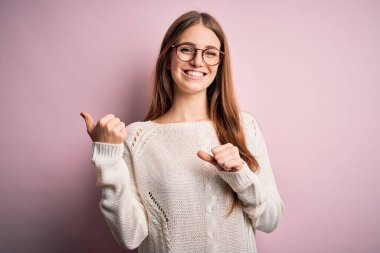 Genç, güzel, kızıl saçlı bir kadın pembe arka planda günlük süveter ve gözlük takıyor.