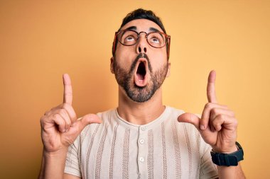 Sarı arka planda günlük tişört ve gözlük takan sakallı genç yakışıklı adam şaşırmış ve şaşırmış bir şekilde parmaklarıyla işaret ediyor ve kollarını kaldırıyordu..