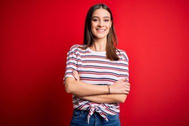 Genç, güzel, esmer, sıradan çizgili tişört giyen, kırmızı arka planda duran mutlu yüzlü, kollarını kavuşturup kameraya bakan bir kadın. Pozitif kişi.