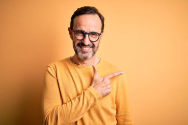 Orta yaşlı, ağarmış, günlük süveter giyen ve gözlüklü, izole edilmiş sarı arka planda neşeli, yüzünde, mutlu ve doğal bir ifadeyle elini ve parmağını gösteren bir gülümseme olan bir adam.