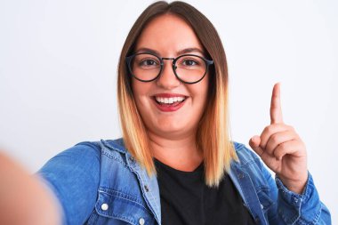 Kot gömlek ve gözlük takan güzel bir kadın, izole edilmiş beyaz arkaplanda bir fikir ya da soru işareti ile şaşırmış bir şekilde selfie çeker.