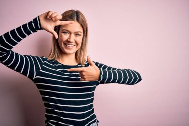 Genç ve güzel sarışın kadın pembe izole edilmiş arka planda sıradan çizgili süveter giyiyor gülümsüyor ve mutlu yüzlü elleri ve parmakları olan bir çerçeve yapıyor. Yaratıcılık ve fotoğrafçılık kavramı.