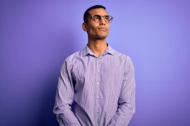 Yakışıklı Afro-Amerikalı, çizgili gömlek ve gözlük takmış mor arka planda gülümseyen ve başka yöne bakan..