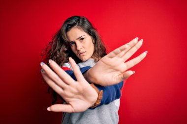 Kıvırcık saçlı genç bir kadın izole edilmiş kırmızı arka planda rahat bir kazak giyiyor. Kollarını kavuşturup, avuç içlerini ters çevirip, kızgın bir yüz ifadesi takınıyor.