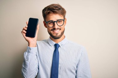 Akıllı telefonu elinde tutan genç bir işadamı mutlu bir yüzü olan, dişlerini gösteren bir gülümsemeyle izole edilmiş beyaz arka planda duruyor.