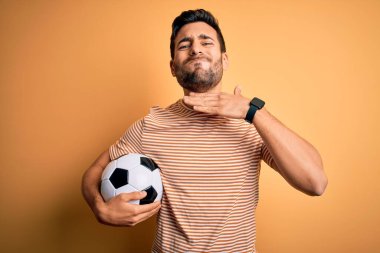 Sakallı yakışıklı oyuncu futbol topu tutarken sarı arka planda boğazını bıçak niyetine kesiyor, saldırganlığı şiddetle tehdit ediyor.