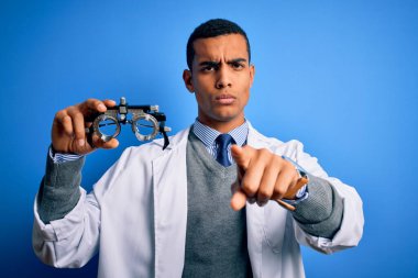 Yakışıklı Afro-Amerikan optik adam elinde optometri gözlükleriyle mavi arkaplanı işaret ederek kamerayı işaret ediyor ve sen, el işareti, ön taraftan pozitif ve kendinden emin bir hareket.