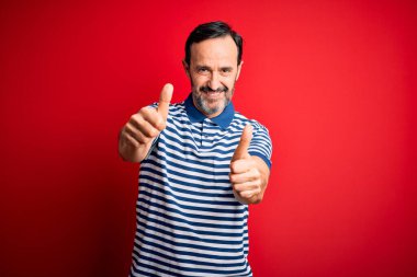 İzole edilmiş kırmızı arka planda dikilen sıradan çizgili polo giyen, el hareketlerini onaylayan, gülümseyen ve başarı için sevinen orta yaşlı beyaz bir adam. Kazanan jest.