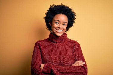 Kıvırcık saçlı, balıkçı yaka kazak giyen, kollarını kavuşturup kameraya gülümseyen güzel Afro-Amerikan kadın. Pozitif kişi.