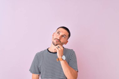 Çizgili tişört giyen genç yakışıklı adam pembe izole edilmiş arka plan üzerinde duruyor ve elleri çenesinde soru sormayı düşünüyor, düşünceli yüz ifadesi. Düşünceli bir yüzle gülümsüyor. Şüphe kavramı.