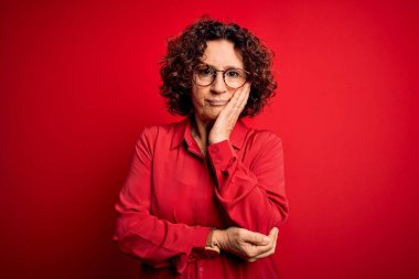 Orta yaşlı, kıvırcık saçlı, rahat gömlekli, kırmızı arka planda gözlüklü güzel bir kadın. Kollarını kavuşturup depresyondan sıkılmış ve yorgun görünüyor..