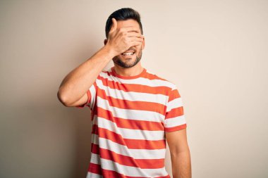 Sıradan çizgili tişört giyen yakışıklı genç adam izole edilmiş beyaz arka planda duruyor. Gülümsüyor ve elleri yüzünde şaşkınlık için gözleri kapatıyordu. Kör kavram.