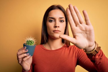 Genç, güzel esmer kadın sarı arka planda küçük bir kaktüs tenceresi tutuyor. Açık elle dur işareti yapıyor. Ciddi ve kendinden emin bir ifadeyle, savunma hareketi yapıyor.