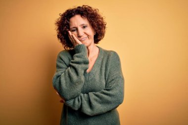 Orta yaşlı güzel kıvırcık saçlı kadın sarı arka planda günlük süveter giyiyor. Kollarını kavuşturup depresyondan sıkılmış ve yorgun görünüyor..