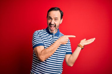 İzole edilmiş kırmızı arka planda dikilen sıradan çizgili polo giyen orta yaşlı beyaz adam kameraya hayret ediyor ve gülümsüyor..