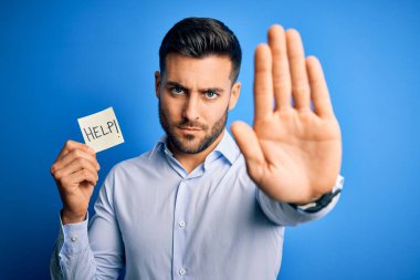 Çok çalışan genç iş adamı mavi arka planda kağıt tutarken yardım istiyor. Elinde dur işareti, ciddi ve kendinden emin bir ifade ve savunma hareketi var.