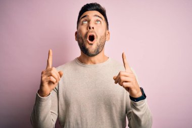 İzole edilmiş pembe arka plan üzerinde duran genç ve yakışıklı bir adam şaşırmış ve şaşırmış bir şekilde yukarı bakıyor ve parmaklarıyla işaret ediyor ve kollarını kaldırıyor..