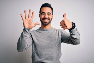 Sakallı, sıradan bir süveter giyen, beyaz arka plan üzerinde duran, altı numaralı parmaklarıyla işaret eden, kendinden emin ve mutlu gülümseyen genç yakışıklı bir adam..