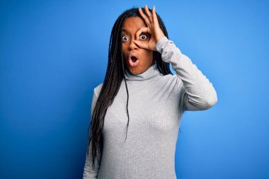 Genç Afro-Amerikalı kadın mavi izole edilmiş arka plan üzerinde günlük balıkçı yaka kazak giyiyor. Şaşırmış yüz ifadesiyle şaşırmış, parmaklarından bakan gözlerle hareket ediyor. İnançsız ifade.