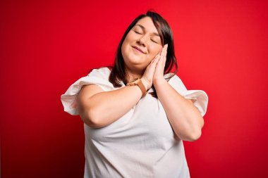 Güzel esmer ve cüsseli bir kadın izole edilmiş kırmızı arka plan üzerinde gündelik tişört giyip uyur, yorgun düşler kurar ve elleri ile birlikte poz verir ve gözleri kapalı gülümserler..