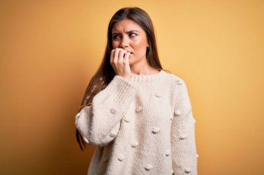 Mavi gözlü genç bir kadın sarı arka planda dikilen sıradan bir süveter giyiyor. Elleri ağızdan tırnak yerken gergin ve stresli görünüyor. Endişe sorunu.